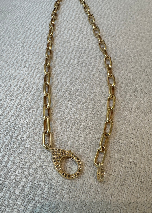 A 17 Link chain with diamond clasp by the woods, featuring large gold vermeil oval links and a circular textured clasp, rests on a textured, light-colored surface.