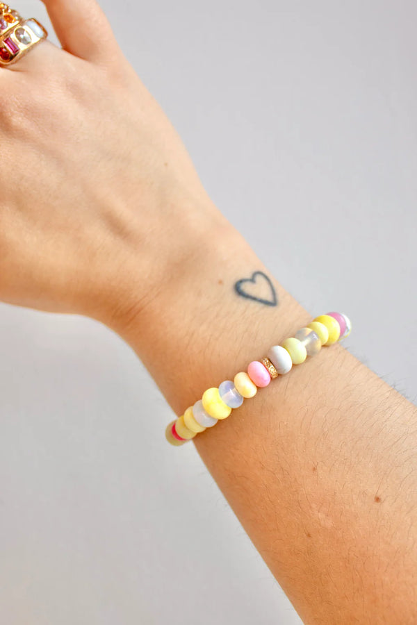 A wrist with a small heart tattoo wears the Scosha Candy Gem Bracelet in Lemonade, featuring pastel beads, along with a gemstone gold ring. The background is plain and light-colored.
