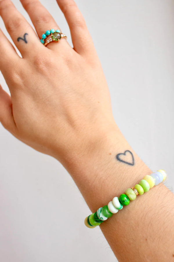 A hand with a heart and Aries tattoo, wearing a gold turquoise ring and the Scosha Candy Gem Bracelet in Cactus, featuring green, white, and yellow gemstone beads. The background is plain and light.