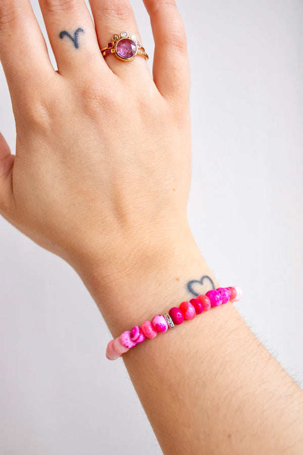 A tattooed hand with a ring wears the Scosha Candy Gem Bracelet in Gelato, featuring pink, red, and white beads of various shapes and shades plus a turquoise cabochon charm. Tattoos shown include a heart and an Aries symbol.