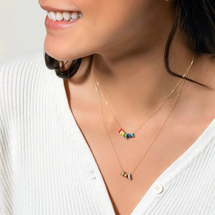 A woman wearing a white buttoned top smiles, showcasing layered gold necklaces. One necklace highlights colorful AR Blue Enamel Beads from Adina Reyter, while the other features a small charm. Her dark hair enhances the casual yet chic look, perfect for any bead party.