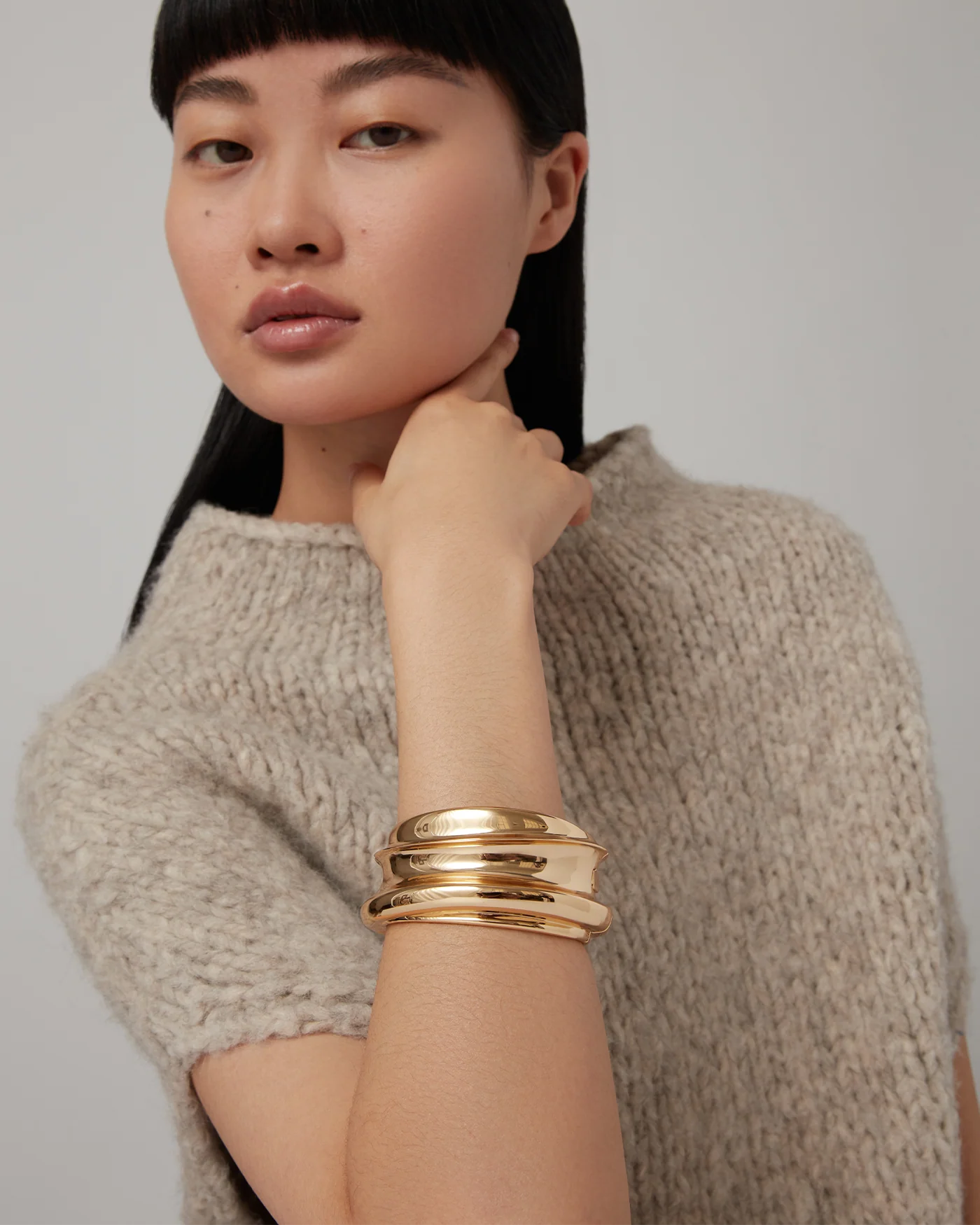 A woman with straight black hair and bangs poses with her hand near her neck, wearing a beige knitted top and the Jenny Bird Mega Lennon Bangle among chunky gold bangles against a neutral background.