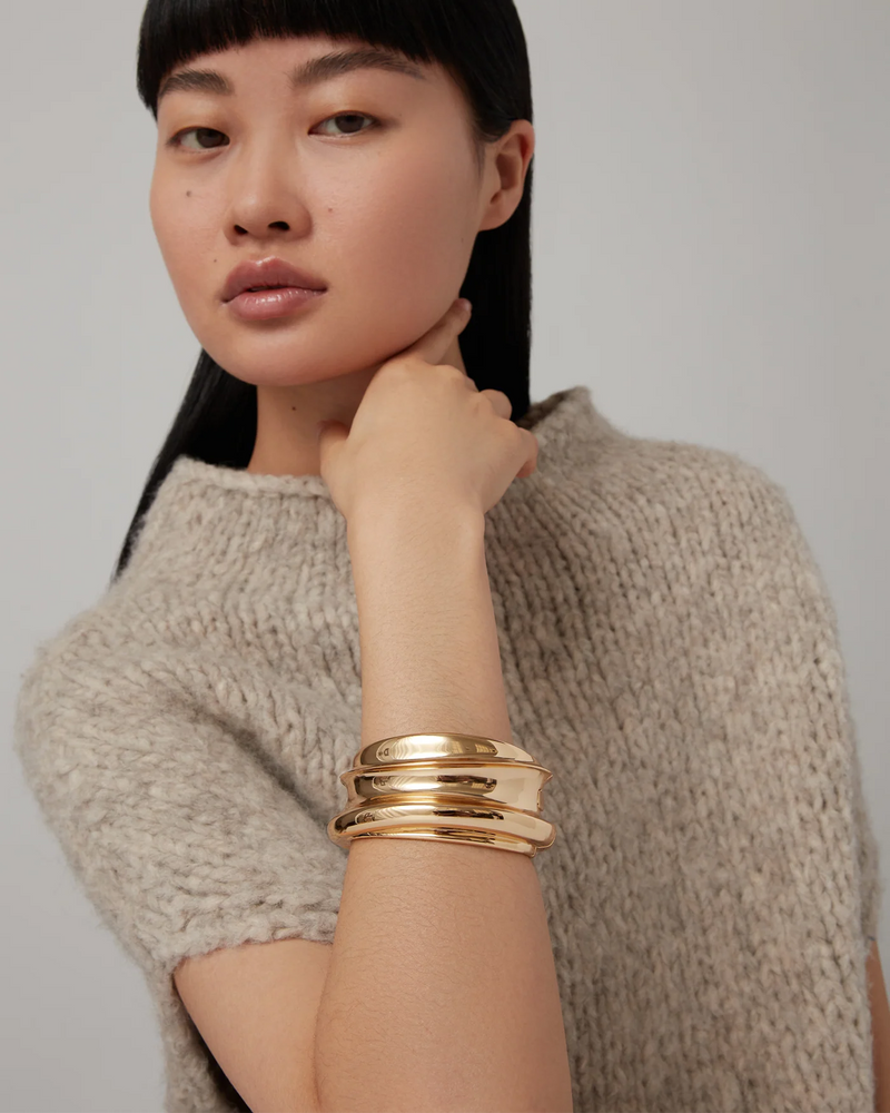 A woman with straight black hair and bangs poses with her hand near her neck, wearing a beige knitted top and the Jenny Bird Mega Lennon Bangle among chunky gold bangles against a neutral background.