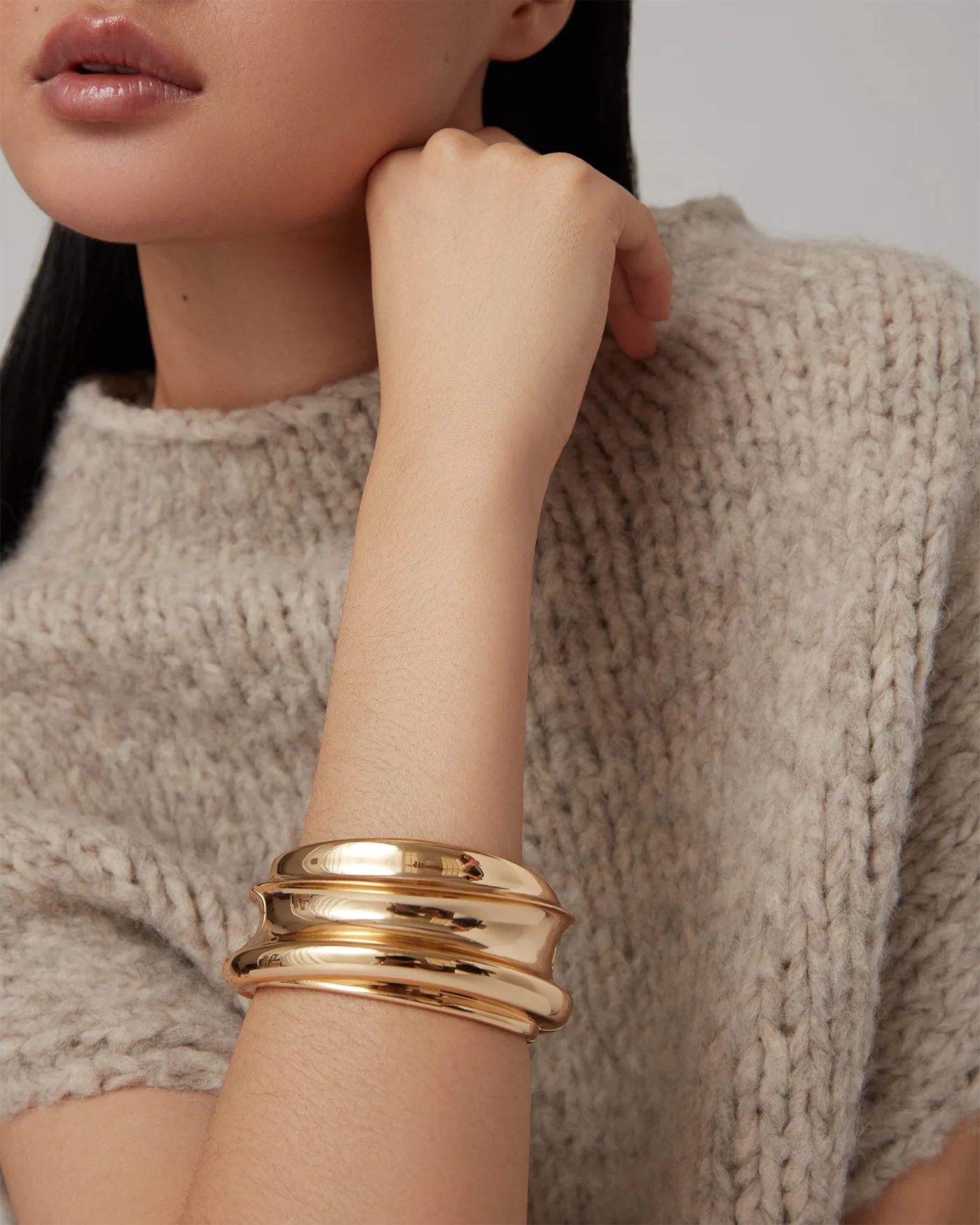 A woman in a chunky beige knit sweater rests her chin on her hand, displaying the Jenny Bird Mega Lennon Bangle and a thick, shiny gold cuff on her wrist. Only the lower half of her face and upper body are visible.