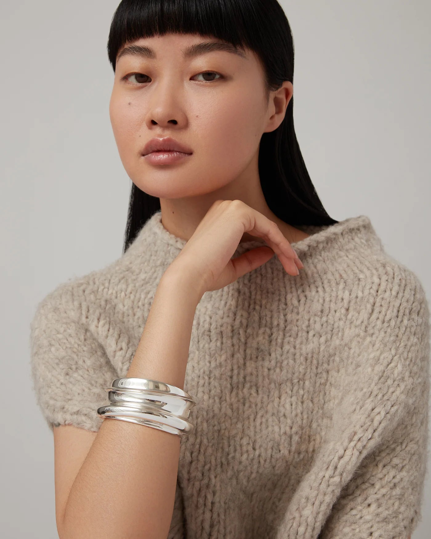 A woman with straight black hair and bangs wears a chunky beige knit sweater and the Jenny Bird Mega Lennon Bangle, stacked with shiny silver bracelets, as she rests her hand near her chin and gazes at the camera.
