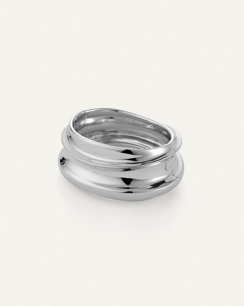 The Jenny Bird Mega Lennon Bangle is a shiny, silver statement piece with a chunky, smooth, wavy design and stacked layers, showcased on a plain white background.