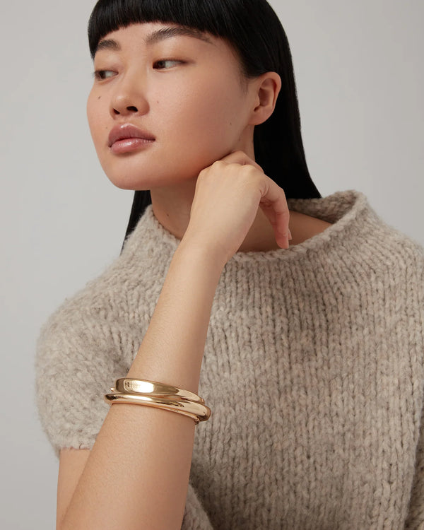 A woman with straight black hair wears a textured beige sweater and gazes to the side, resting her chin on her hand, while showcasing two Jenny Bird Lennon Bangles on her wrist against a plain, light background.