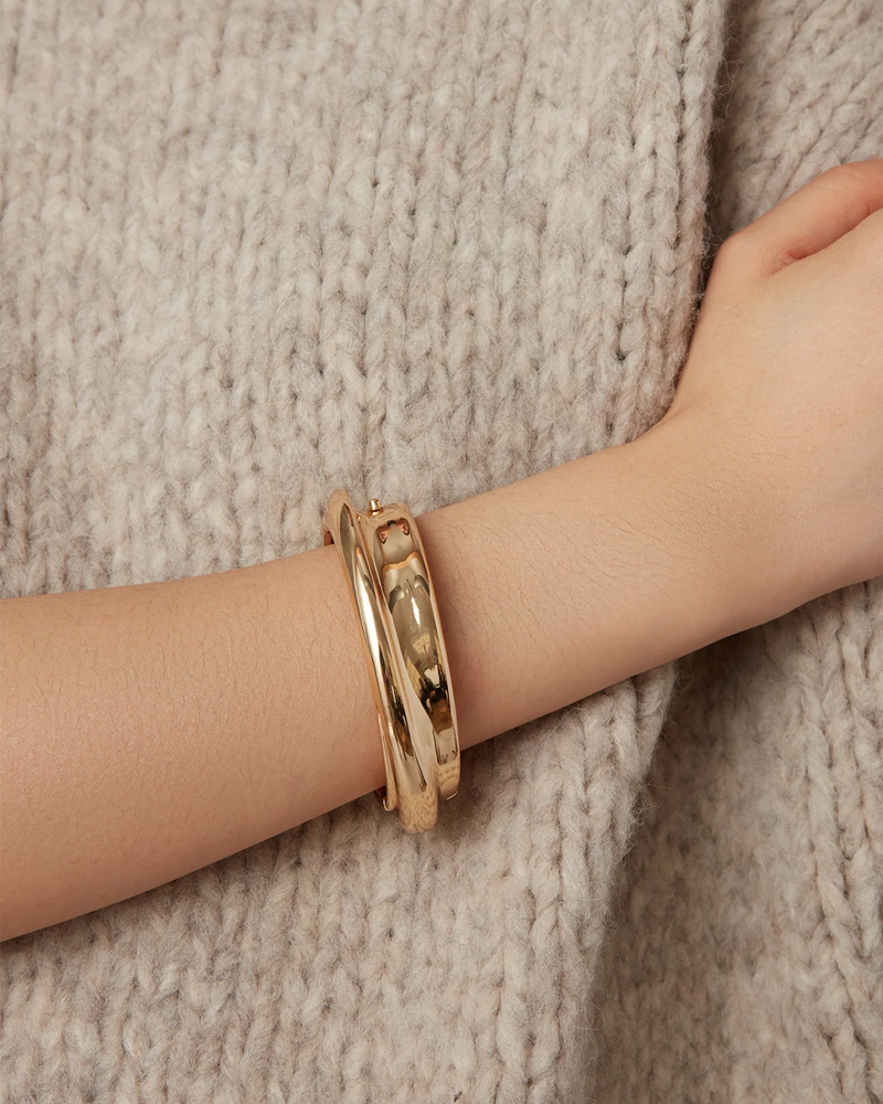 A close-up of a person’s arm shows two thick, shiny Lennon Bangles by Jenny Bird in gold tone against chunky beige knit fabric.