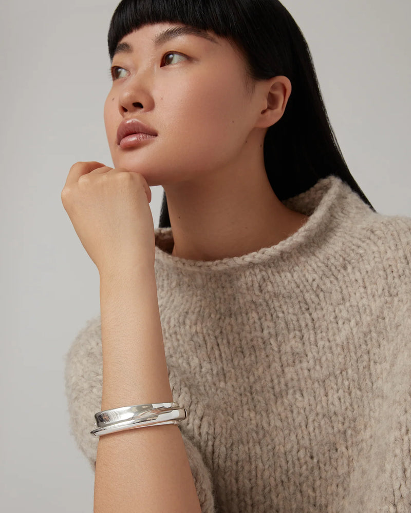 A woman with straight black hair and blunt bangs wears a chunky beige knit sweater and the Lennon Bangle by Jenny Bird. She thoughtfully rests her chin on her hand, gazing upward to the left against a plain background.