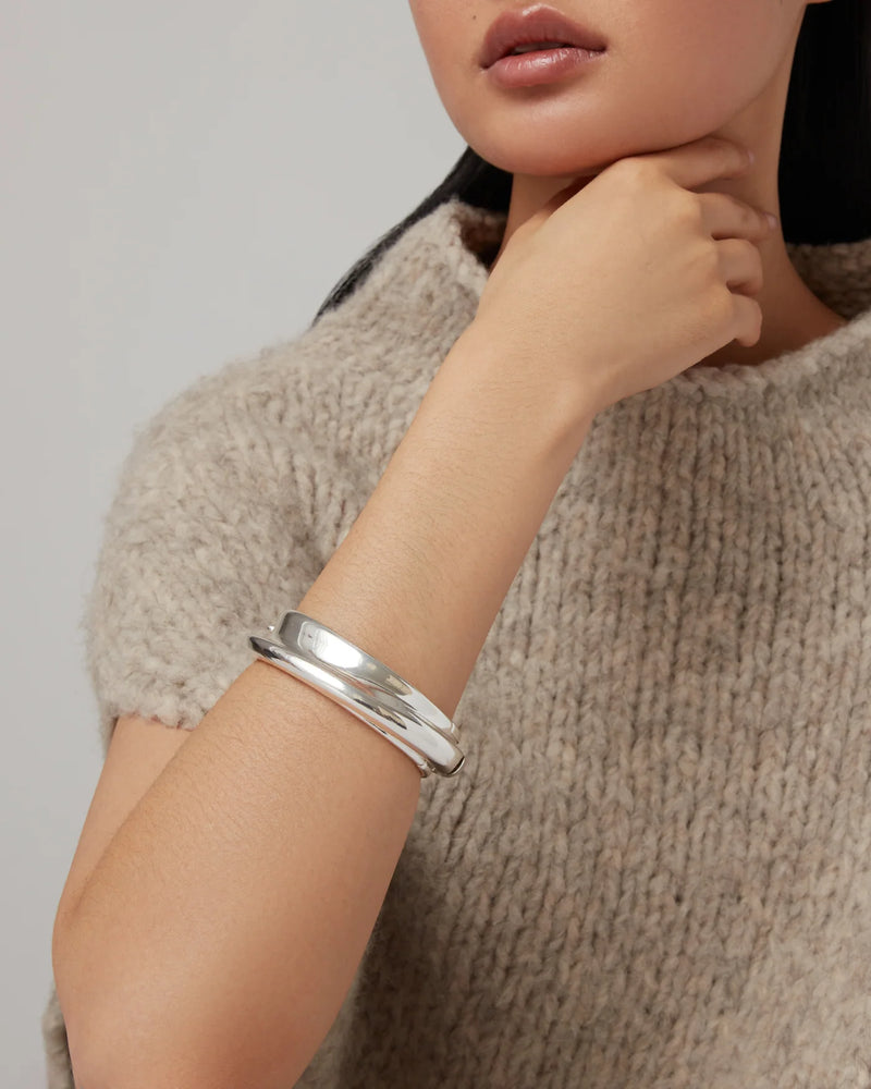 A woman in a beige knit top rests her chin on her hand, highlighting the shiny Jenny Bird Lennon Bangle on her wrist. The image is cropped to show only her lower face and upper torso.