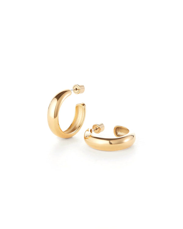 The Staple Hoop Earrings - Medium by Jenny Bird are shown on a white background. One medium-sized, thick 14k gold hoop earring stands upright, while the other lies flat to reveal its post and clasp. Both feature smooth, shiny surfaces.
