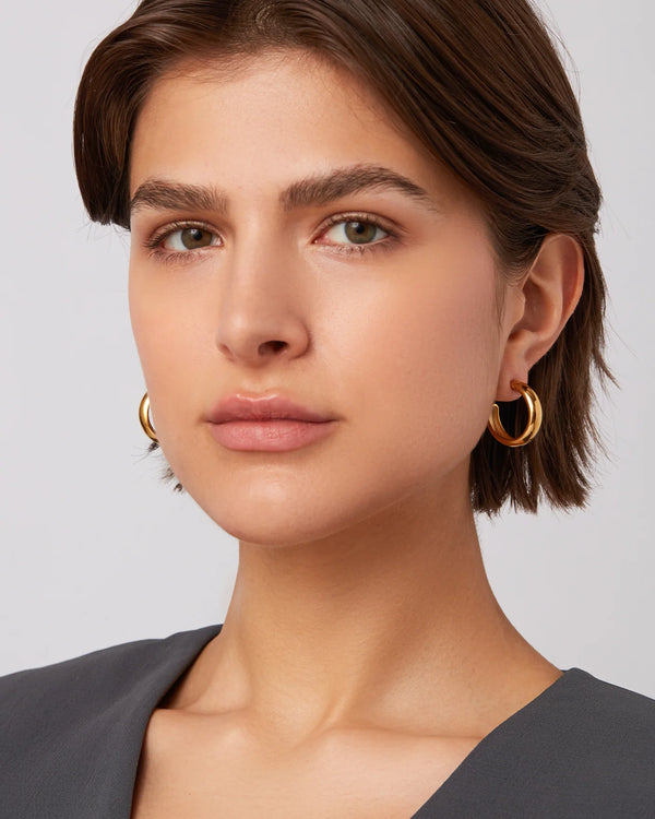 A woman with short brown hair wears Jenny Bird Staple Hoop Earrings - Medium in lightweight 14k gold. She gazes neutrally at the camera, dressed in a dark grey top, natural makeup, and stands against a plain light background.