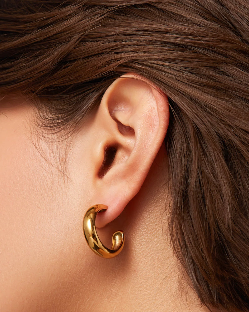 A close-up of a persons ear with straight brown hair, wearing Jenny Bird Staple Hoop Earrings - Medium in shiny 14k gold, featuring a thick yet lightweight design.