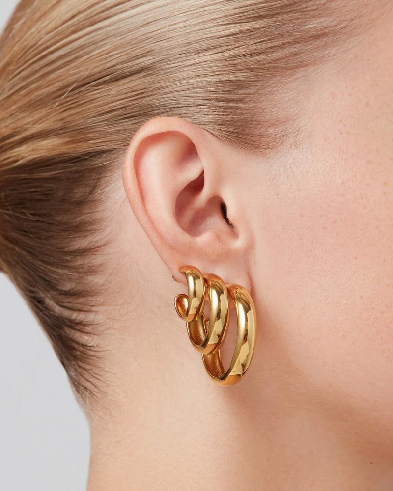 Close-up of a persons ear wearing Jenny Bird Staple Hoop Earrings - Medium, a set of three chunky, interlocking 14k gold hoops. The lightweight earrings stand out against the neutral background; the person has light skin and pulled-back blonde hair.