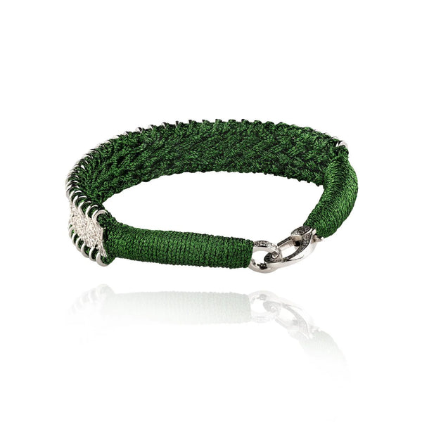 The Janeiro Grass bracelet by Van Den Abeele is a handmade green woven piece with a silver metal clasp, featuring intricate detailing and a textured design. Its displayed on a white background, casting a subtle reflection below it.