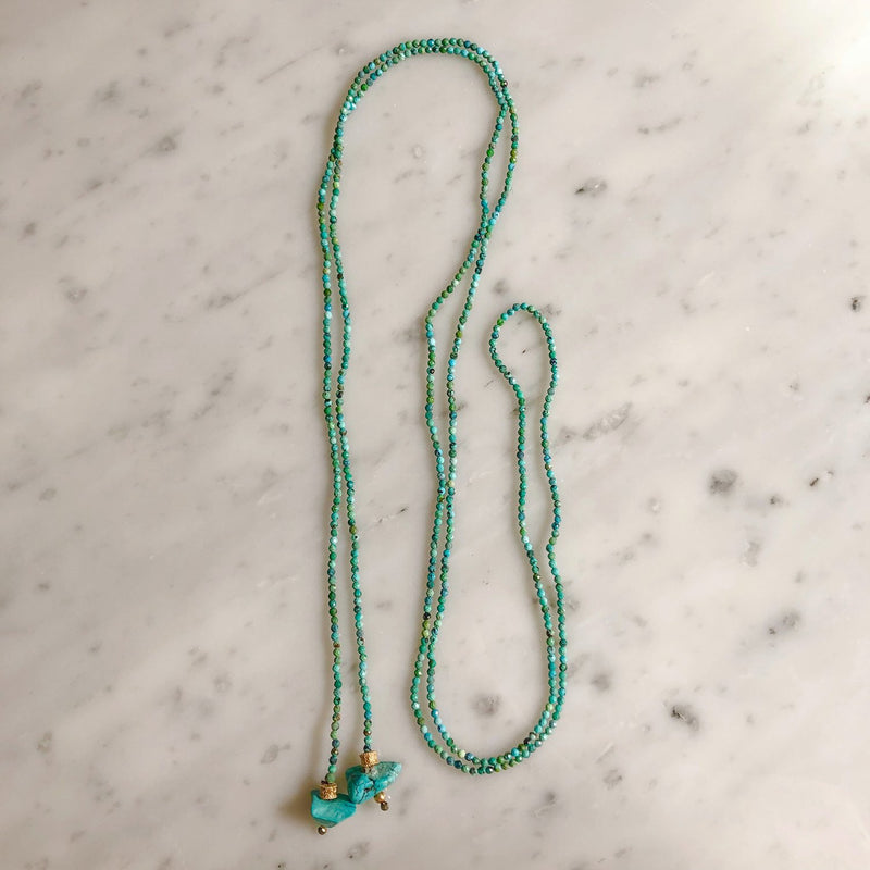 The KEVIN Lariat by Bohemian Rhapsodie Paris, a long turquoise necklace with intricate beadwork and gold-plated detailing, features a pendant with two stones and small gold accents. It is elegantly arranged on a light marble surface.
