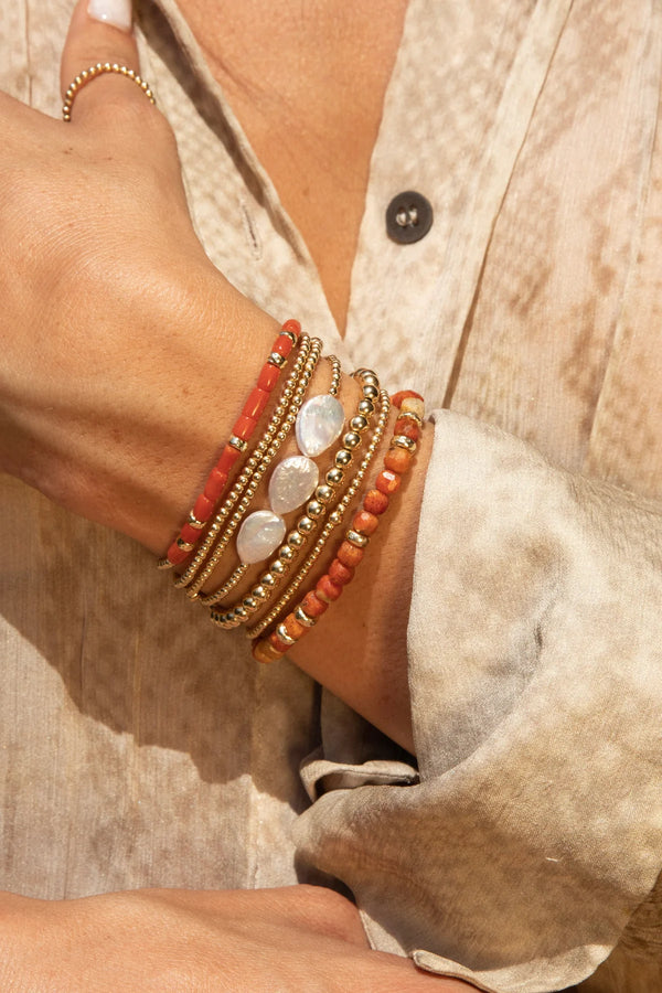 A persons arm showcases the Karen Lazar BARREL CORAL AND HAMMERED RONDELLE PATTERN BRACELET with gold beads, orange stones, and white pearls, complemented by a beige textured button-up shirt. Their hand touches a wrist adorned with a gold ring featuring barrel coral gemstones.