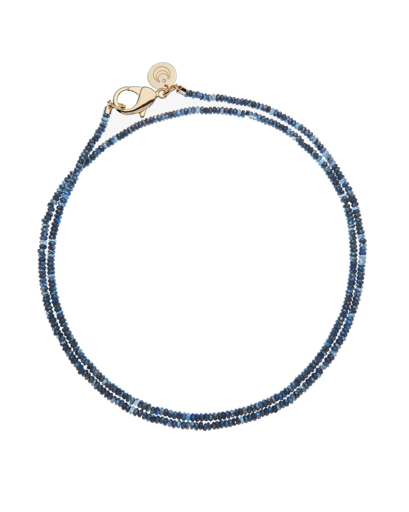 The Jane Win Diamond Cut Double Wrap Beaded Necklace in Kyanite features small, faceted beads, a gold clasp, and a round gold charm, shown arranged circularly on a white background.