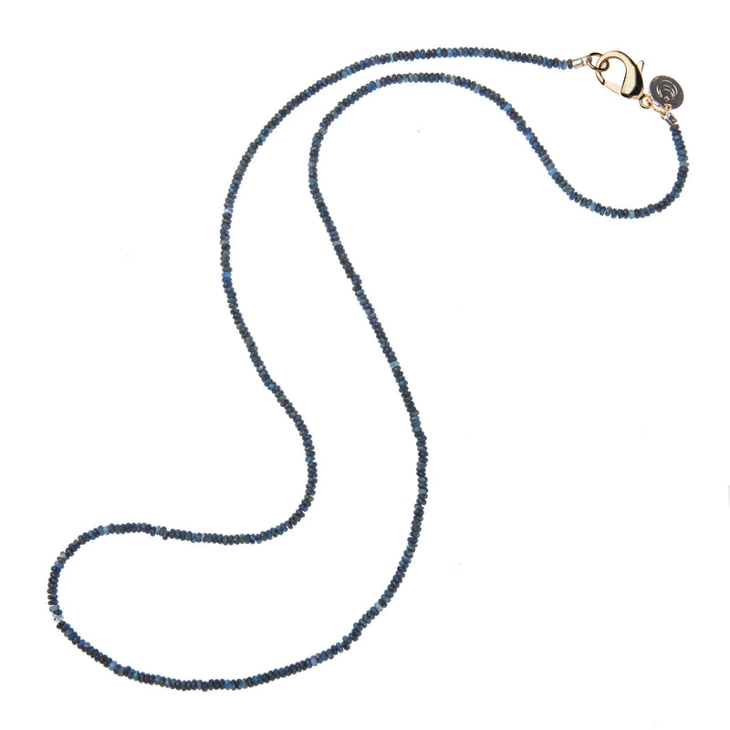 The Jane Win Diamond Cut Double Wrap Beaded Necklace in Kyanite features small, round dark blue beads, a gold clasp, and a round charm near the clasp, shown on a white background.