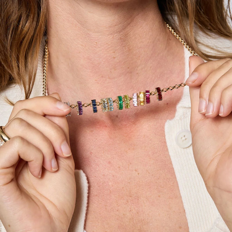 A person in a white top holds a stunning 14k gold Adina Reyter necklace adorned with the Emerald Baguette Big Bead and colorful gemstone pendants like purple, blue, pink, and clear stones. Hands emphasize the exquisite birthstone necklace.