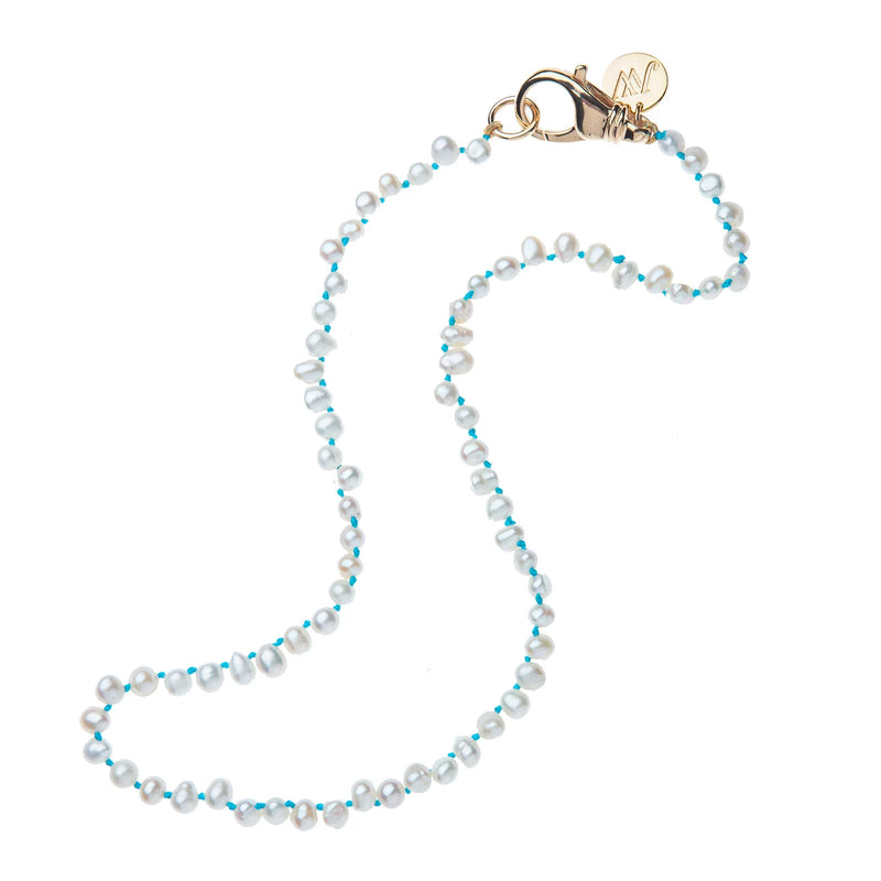 The Mermaids Gift Pearl Necklace by Jane Win features small white pearls, tiny blue semi-precious stones, and a 14k gold-plated brass lobster clasp with a round gold charm, displayed in a loose curve on a white background.