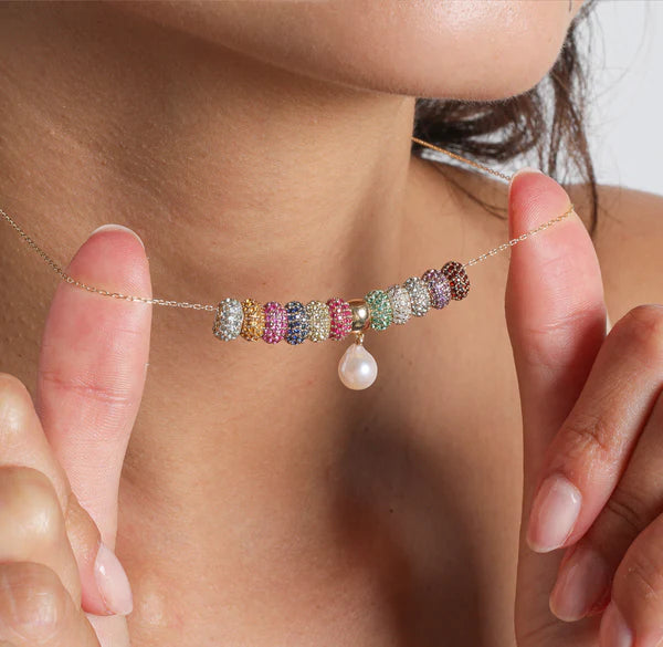 A close-up view captures an individual delicately holding the Wide Pavé Blue Sapphire Mini Bead necklace by Adina Reyter, with colorful beaded rings and a dangling pearl pendant. Their fingers gently touch the 14k gold chain, highlighting the elegant design against their skin, complemented by a radiant amethyst birthstone.