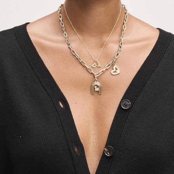 An individual wearing a black cardigan with undone buttons showcases layered necklaces, featuring a heart shape and lock pendant on a chunky chain. Completing the stylish and minimalist look is the Groovy Diamond Initial Charm by Adina Reyter, adorning a delicate chain in 14k yellow gold.