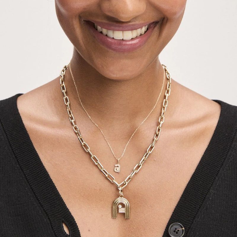 A person wearing two layered 9K yellow gold necklaces smiles softly. The shorter piece boasts a small rectangular pendant, while the longer one showcases an arch-shaped design. Complementing their black top, the Groovy Diamond Initial Mini Bead Charm by Adina Reyter adds a touch of whimsy to this elegant ensemble.