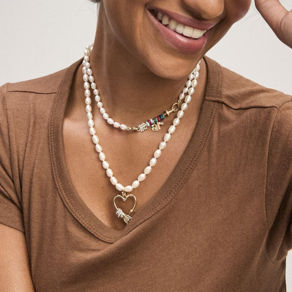 A woman in a brown V-neck shirt smiles, wearing layered pearl necklaces—one with a 14k yellow gold heart pendant and another featuring Adina Reyters Diamond Arrow Hinged Charm for Mini Beads.