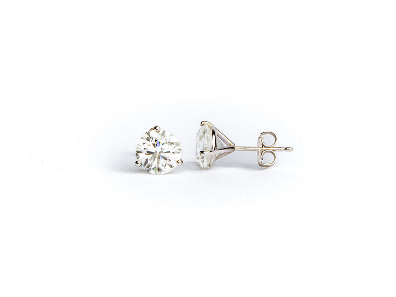 The Monarch Market Lab Grown Studs feature pear-shaped diamonds in a simple silver prong setting. One earring faces upright, revealing the diamonds front, while the other lies on its side, showing the back and post. Both rest on a white surface.