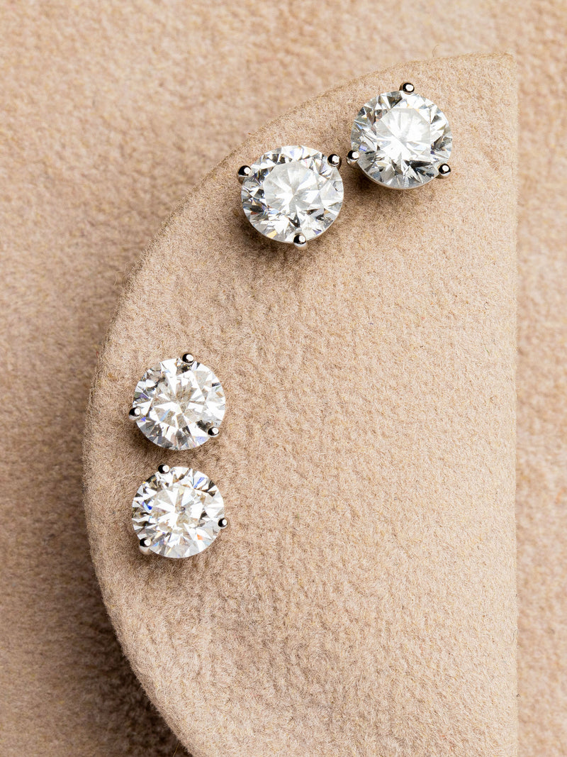 Three Lab Grown Studs by Monarch Market, each pair featuring round diamonds in minimalist four-prong white metal settings, are showcased on a soft, textured light brown fabric.