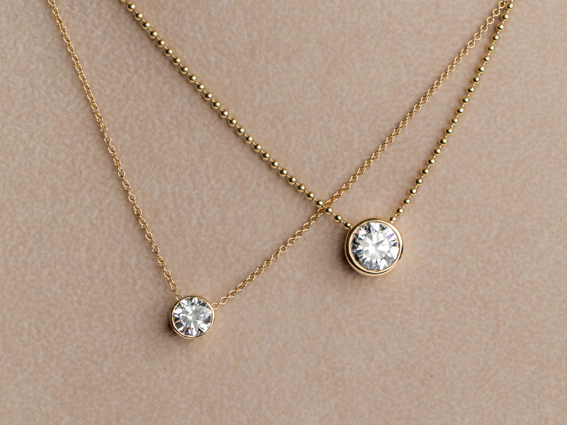 Two delicate Monarch Market necklaces on a soft beige background; both feature lab-grown diamond pendants. The Lab Grown Small Gold Bezel Necklace is elegantly positioned next to a larger pendant, showcasing simplicity and elegance.