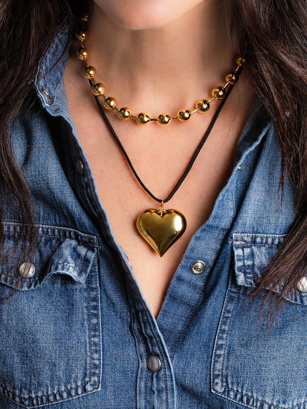 A woman in a blue denim shirt wears handmade jewelry: an Oiya Gold Heart Suede Necklace with a shiny gold heart pendant, and a gold beaded necklace. Only the lower part of her face is visible.