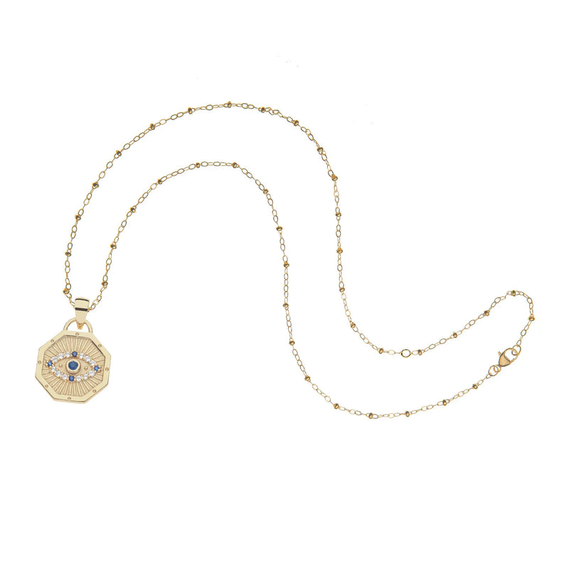 The Jane Win PROTECT Evil Eye Petite Embellished Pendant is a limited edition gold necklace with a delicate chain, featuring an octagonal evil eye pendant with a blue center and surrounding white stones.