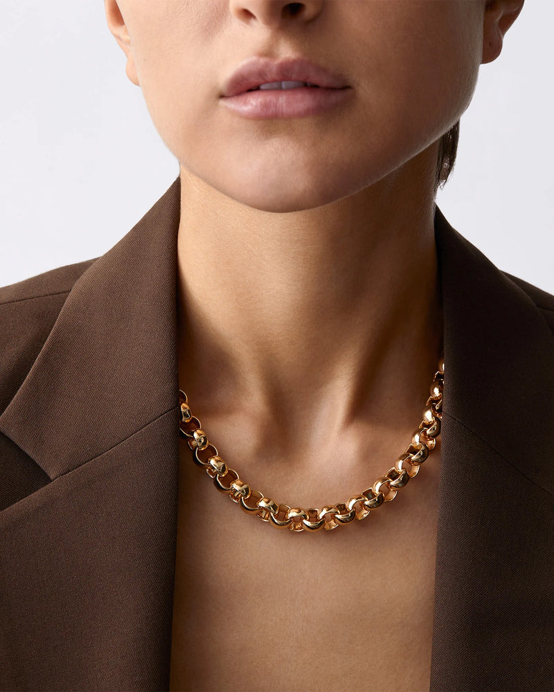 Someone wearing the Jenny Bird Rodin Chain, a chunky, high-polish gold chain necklace, paired with a brown blazer. The image precisely highlights the neck and shoulders against a neutral background.