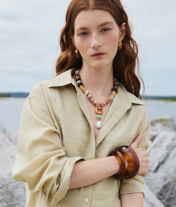 A woman with long red hair stands outdoors by rocks and water, wearing a beige button-up shirt, gold earrings, chunky necklace, and the Lizzie Fortunato Ridge Cuff in Tortoise layered with large brown bangles.