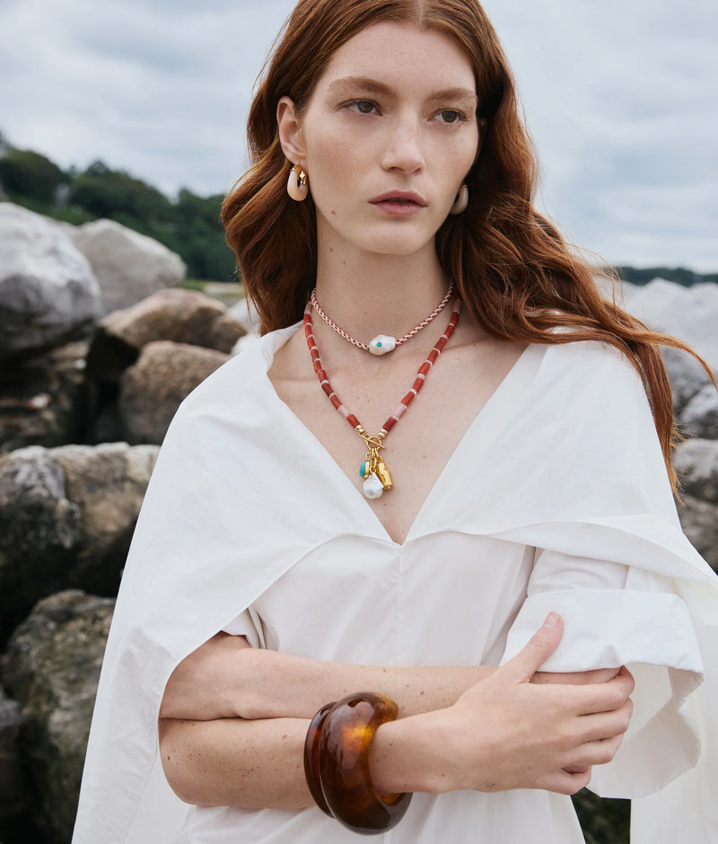 A woman with long red hair stands outdoors by large rocks, wearing a white blouse and the Ridge Cuff in Tortoise by Lizzie Fortunato, along with layered pearl and coral necklaces and gold hoop earrings, gazing thoughtfully into the distance.
