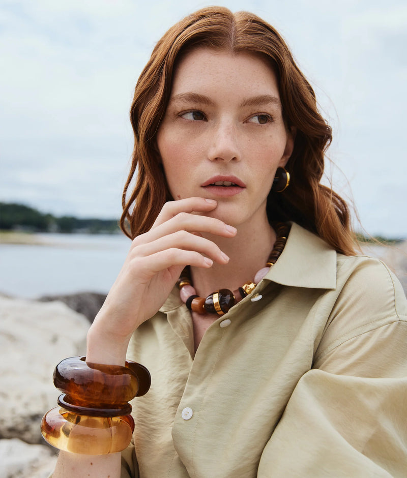 A woman with long red hair sits outdoors on a sandy beach, wearing a beige shirt and the Ridge Cuff in Tortoise by Lizzie Fortunato. She gazes thoughtfully to the side with one hand near her face.