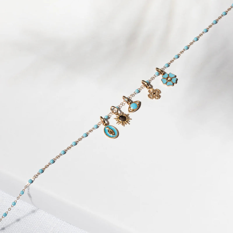 The Sun Pendant by Gigi Clozeau is a delicate 18k gold chain bracelet with turquoise beads and five gold charms, including a sun, an eye, and a flower, displayed diagonally on a light background.