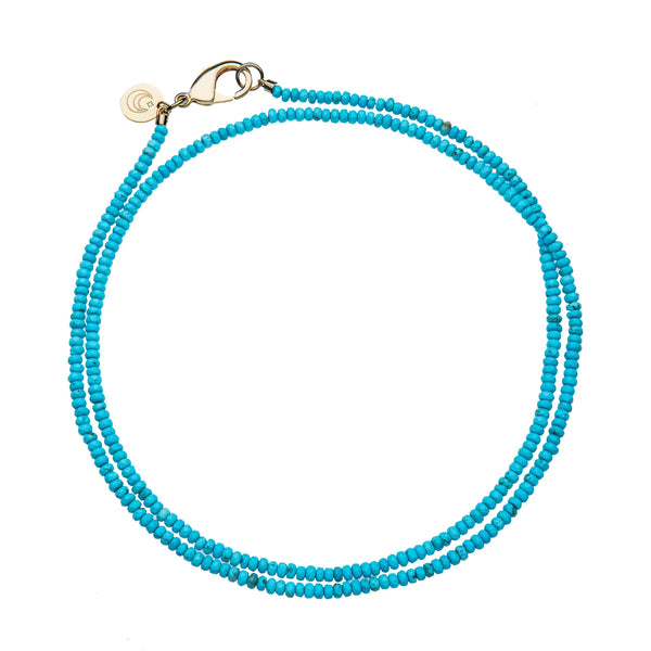 The Jane Win Double Wrap Diamond Cut Beaded Necklace in Turquoise features small, bright beads, a 14k gold-plated clasp, and a round charm. Its displayed in a circular shape on a white background.