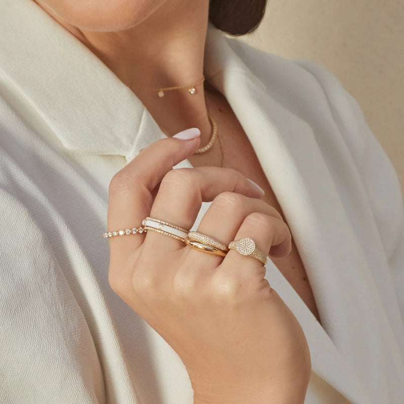 A woman in a white outfit elegantly displays her hand adorned with multiple 14k gold rings, featuring the 3 Diamond Enamel Stack Ring White by ef collection and a heart-shaped design. She wears a delicate gold necklace and bracelet, creating a sophisticated look against a neutral backdrop.