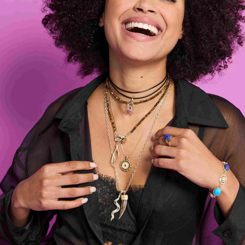 A smiling woman with curly hair models the Jane Win Double Wrap Diamond Cut Beaded Necklace in Tiger's Eye, styled with a sheer black blouse, statement ring, and blue gemstone bracelet against a vivid pink background.