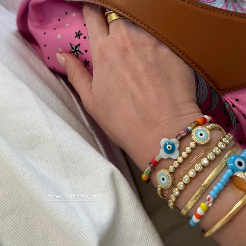 Hand adorned with Aaron Bashas Womens Flower Eye Beaded Stretch Bracelet featuring eye motifs and gold accents rests on white cloth. Partially visible: a tan bag with brown handle and pink star-patterned fabric, alongside a handcrafted token.