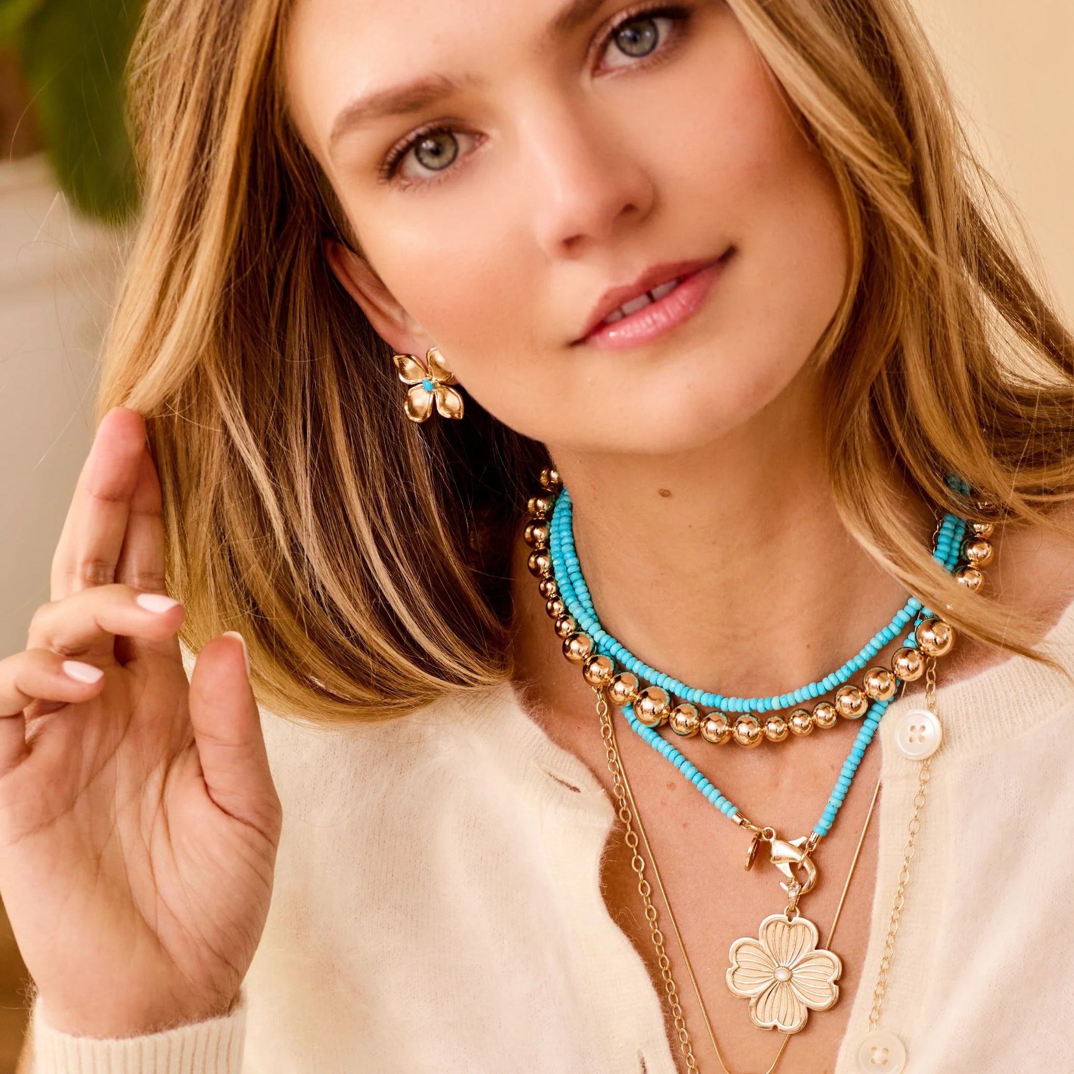 A woman with long blonde hair wears the Sun Reflection Beaded Necklace by Jane Win, featuring gold and turquoise layers. Paired with a cream cardigan, she displays a floral earring and a relaxed, natural expression as she gently touches her hair.