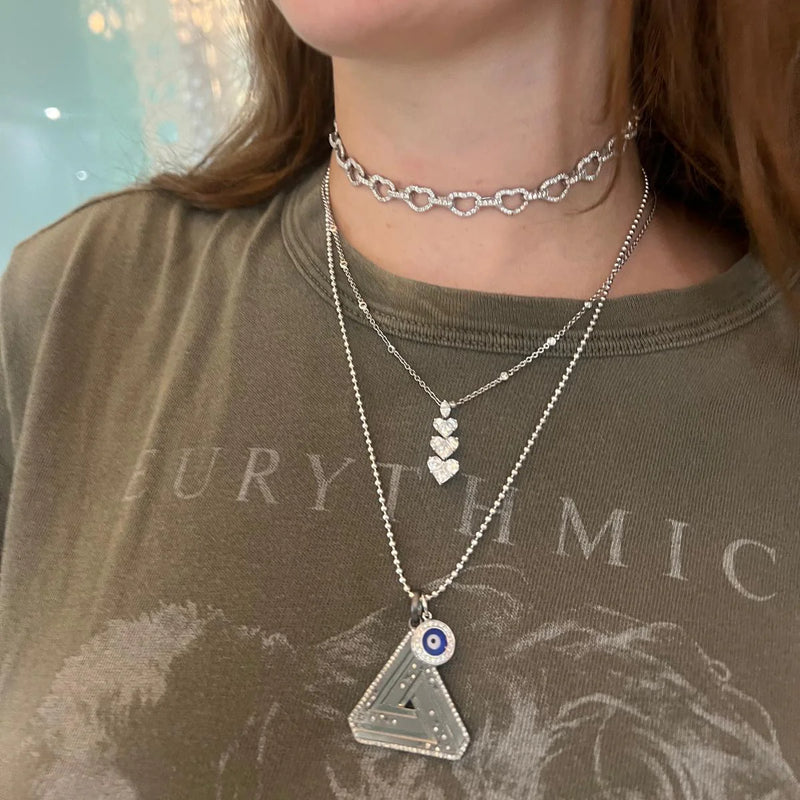 Wearing a trio of striking silver necklaces, including a chain link choker, heart pendant necklace, and Aaron Bashas empowering Large Abracadabra Triangle Series 5 with an eye design. They sport a dark green t-shirt with partially visible text.