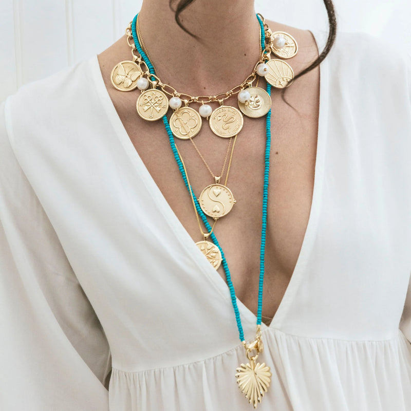A woman in a white deep V-neck dress wears layered gold coin necklaces, including Jane Wins Double Wrap Diamond Cut Beaded Necklace in Turquoise, with 14k gold plating, turquoise beads, diamond cut beads, an engraved heart pendant, and pearl accents.