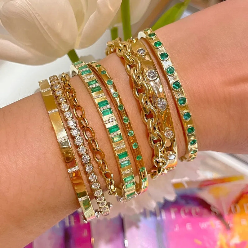 A wrist showcases a collection of gold and sterling silver bangles with diverse designs, such as gems, diamonds, and links. A standout is the Two-Toned Flip Colored Tourmaline Bangle. In the back, blurred flowers and a colorful surface appear. Includes Three Stories Jewelrys Mixed Metal Plain Diamond Bangle for added elegance.