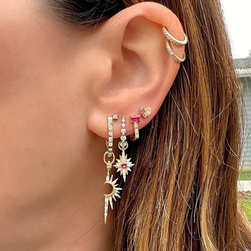 Close-up of a person’s ear adorned with multiple earrings, featuring Three Stories Jewelrys Small Shooting Star Charms in 14K yellow gold, sun, moon, and heart studs. A diamond accent adds sparkle, while two cuffs enhance the upper ear. Light brown hair frames the scene beautifully.
