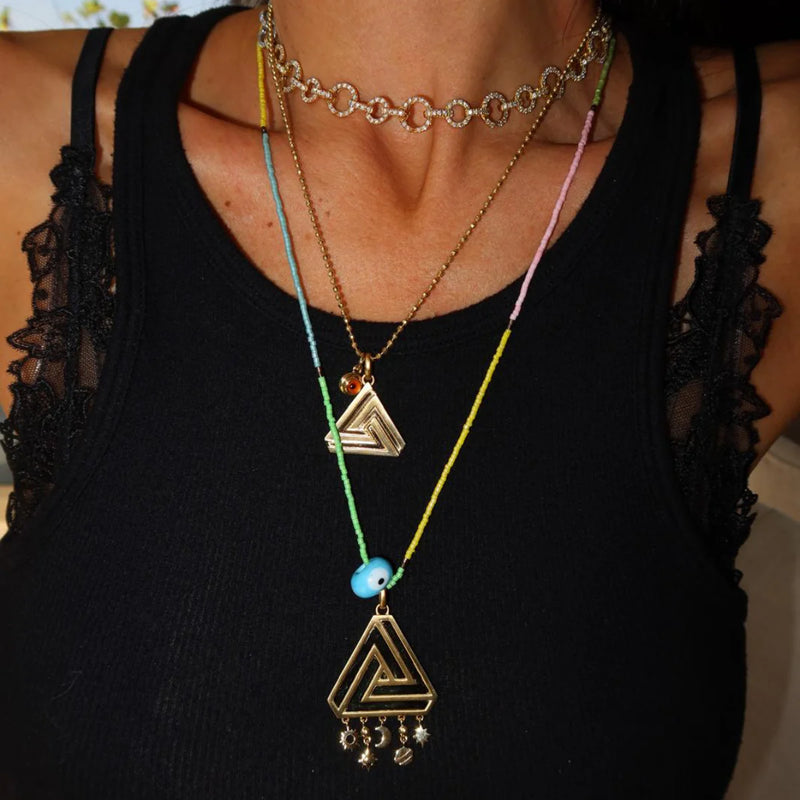 Close-up of a person in a black lace-trimmed tank top, adorned with necklaces. The gold choker with chain links symbolizes self-belief, while others have Aaron Bashas Small Abracadabra Triangle pendants. A blue bead piece hangs from colorful strings, representing personal empowerment.