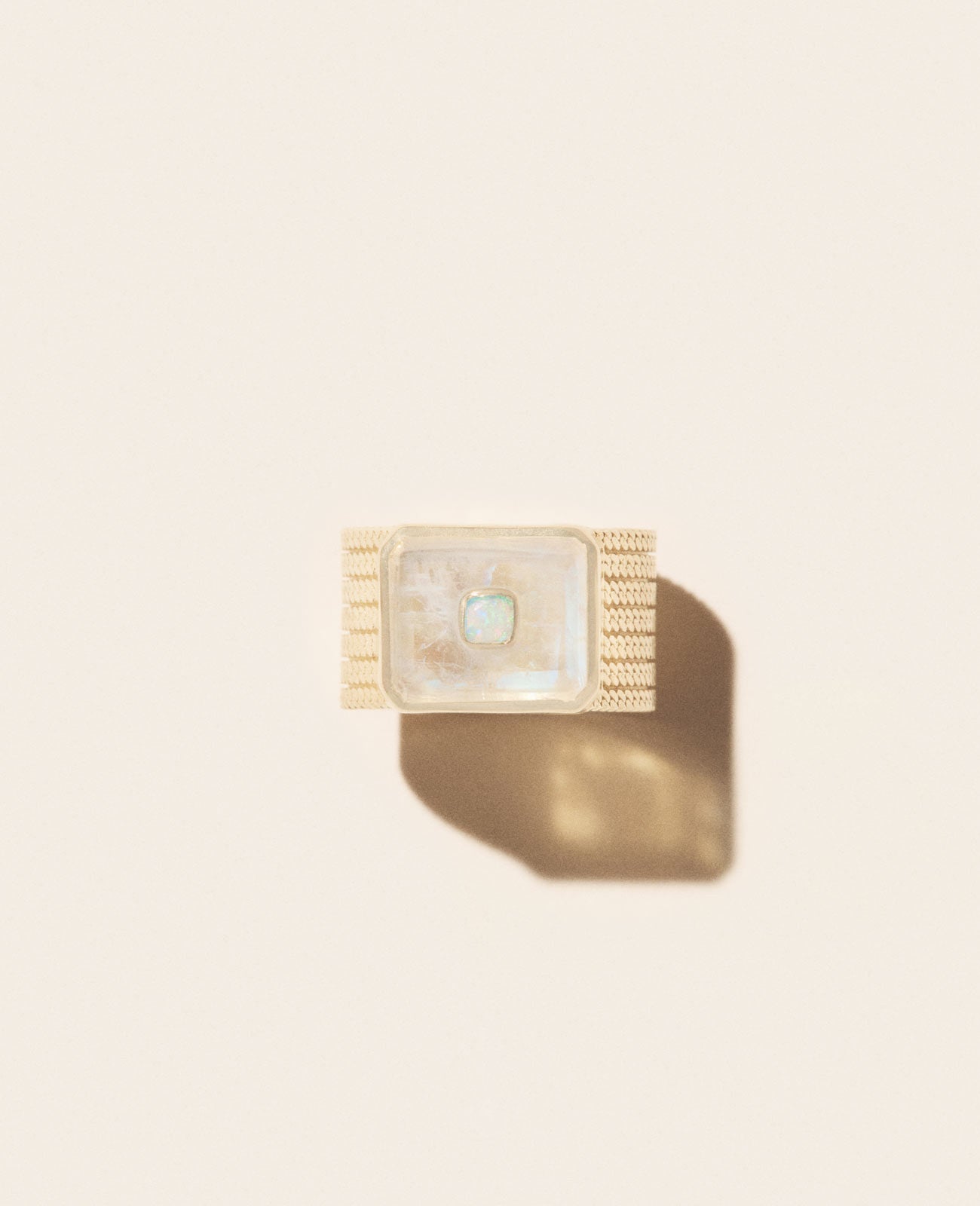 Placed on a light beige background, the Pascale Monvoisin ANTON N°2 MOONSTONE Ring features an openworked textured band in yellow gold with a square, translucent center stone casting an intriguing shadow.